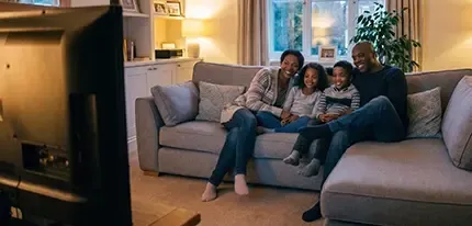 A family of four cuddles together on a couch watching television at night.